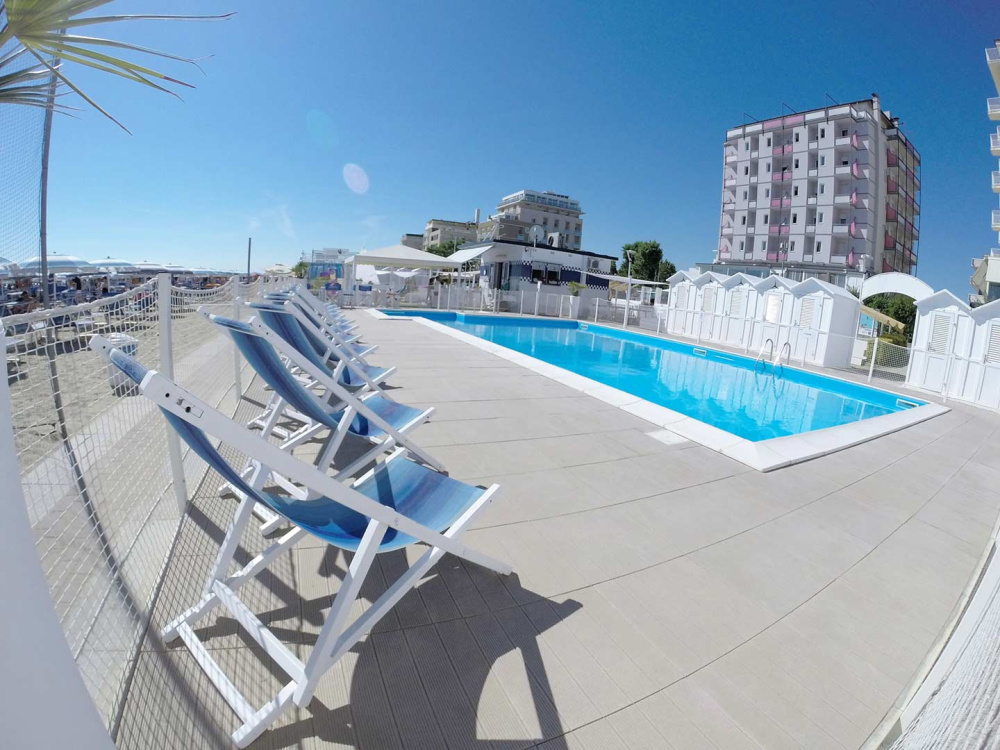 Piscina all'aperto con lettini blu, vicino alla spiaggia e palazzi sullo sfondo.