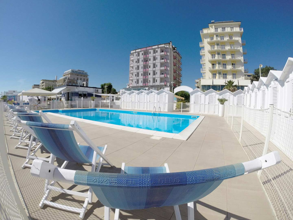 Piscina all'aperto con lettini e cabine bianche, cielo sereno.