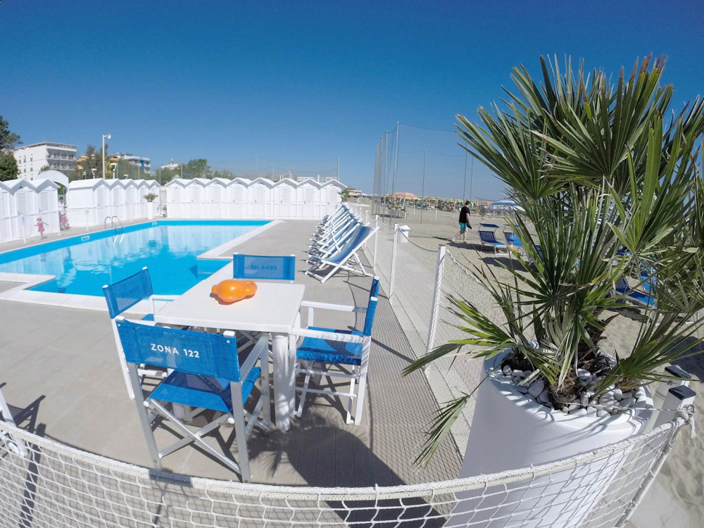Piscina all'aperto con sedie blu, tavolo e palme su una spiaggia assolata.